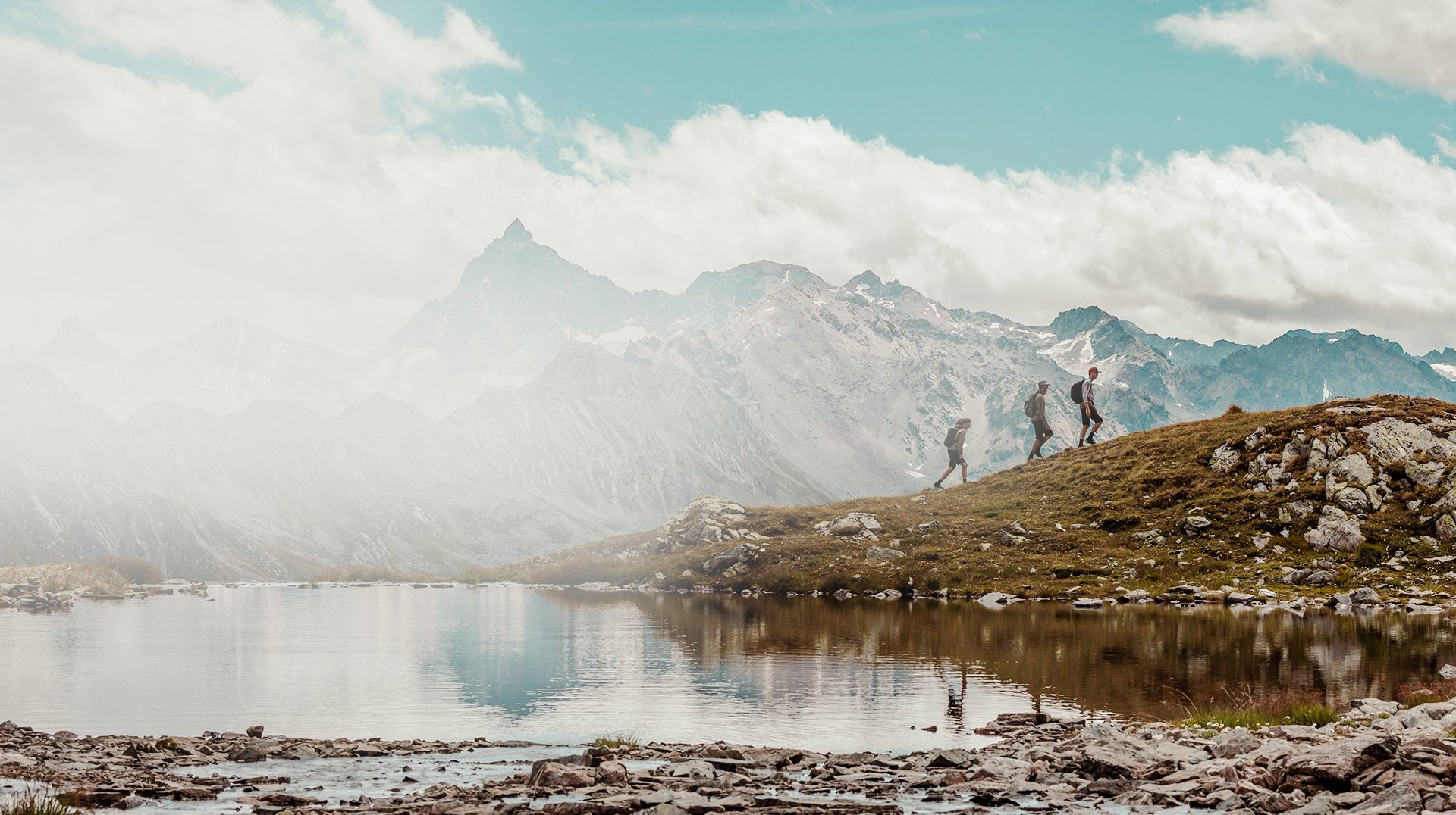Wanderer auf einem Bergpfad mit spiegelndem See und Alpenkulisse